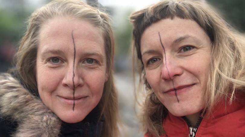 Zwei Gesichter von Menschen, die eng nebeneinander stehen. Über beide Gesichter läuft je von der Stirn bis zum Kinn ein dünner schwarzer Strich.